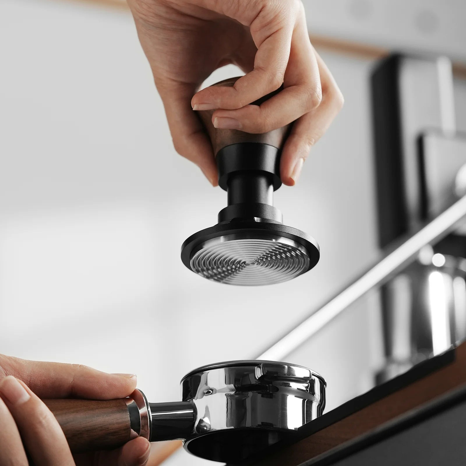 Close-up of hands using the black MHW-3BOMBER Armor Series Coffee Tamper 2.0, featuring adjustable tamping depth, to press ground coffee into a portafilter in a modern kitchen, showcasing the precision and consistency of the coffee-making process.