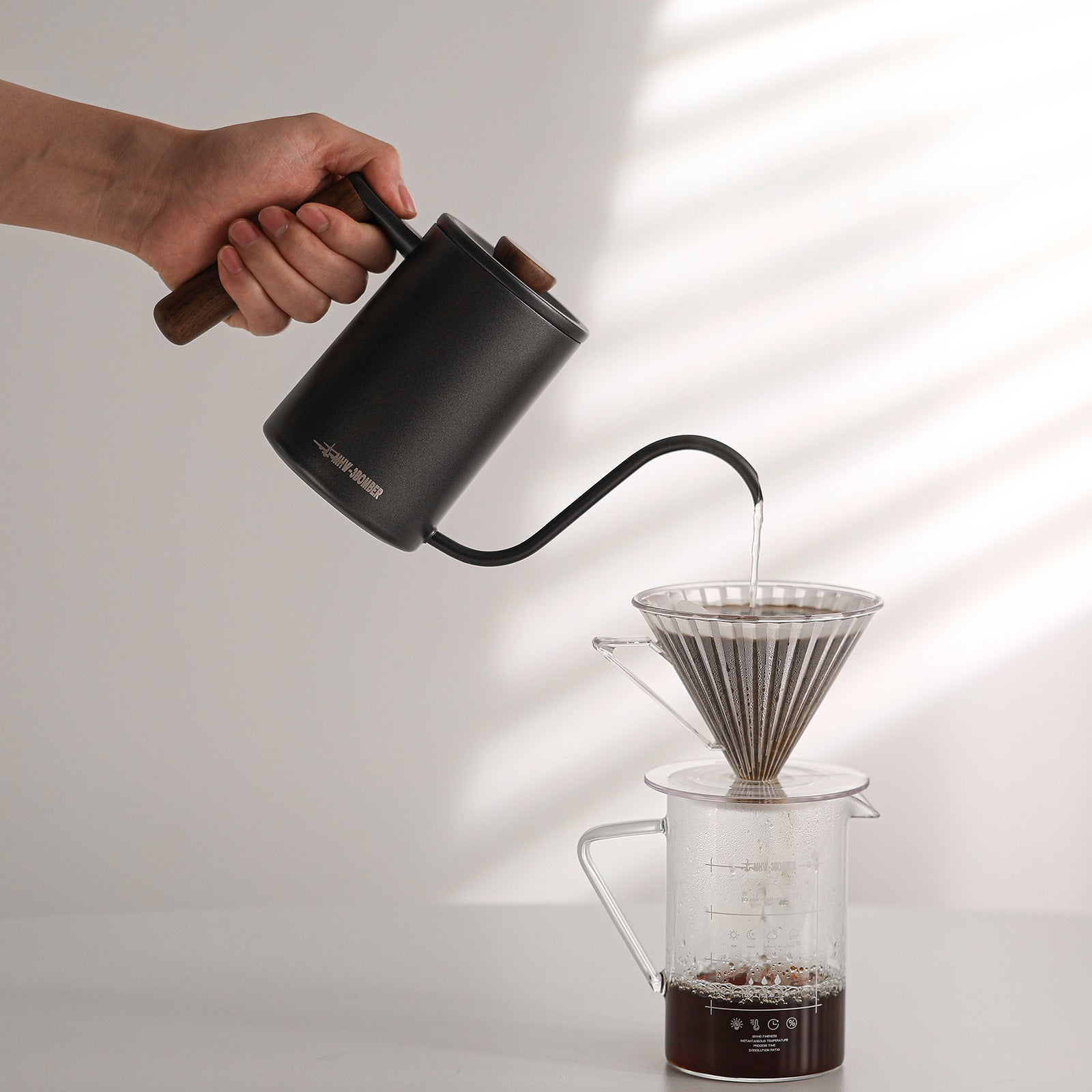 A hand pours hot water from a sleek MHW-3BOMBER Planet Hand Brewing Kettle into a metal coffee dripper on a glass carafe. The setup rests on a light surface with a neutral backdrop, and steam swirls as the coffee brews below.