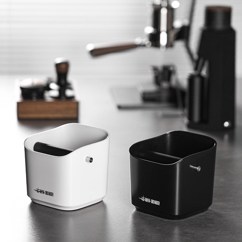 On a kitchen counter, two MHW-3BOMBER Coffee Espresso Square Knock Boxes—one white and one black—stand ready. With a durable cylindrical design featuring an open top and central rod, they are easy to clean. The coffee machine fades into the background, highlighting their functional elegance.