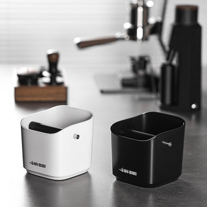 On a kitchen counter, two MHW-3BOMBER Coffee Espresso Square Knock Boxes—one white and one black—stand ready. With a durable cylindrical design featuring an open top and central rod, they are easy to clean. The coffee machine fades into the background, highlighting their functional elegance.