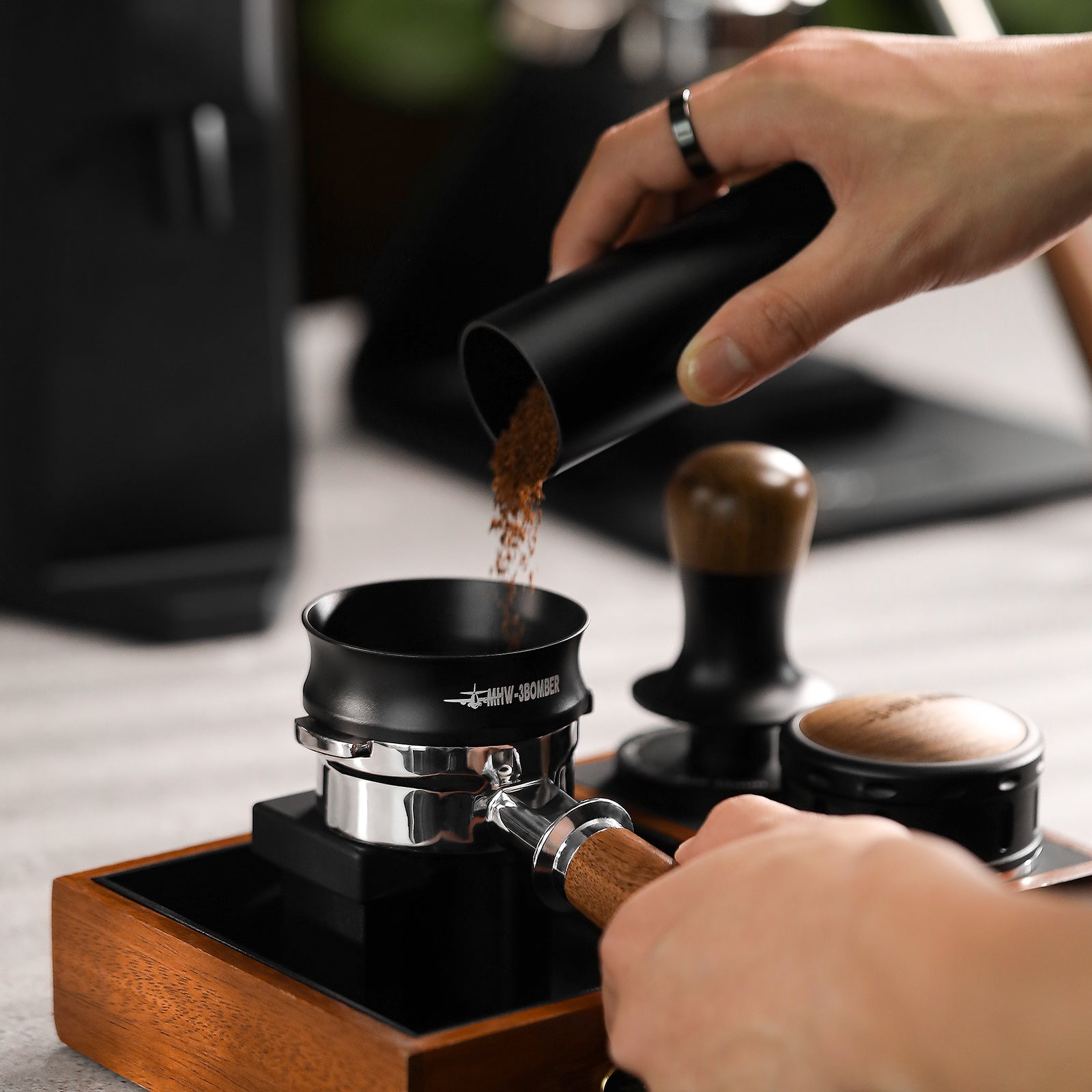 A close-up shows hands preparing coffee using the MHW-3BOMBER Espresso Magnetic Coffee Dosing Funnel to pour ground coffee into an espresso machine accessory, with a tamper and wooden stand nearby on a modern kitchen counter.