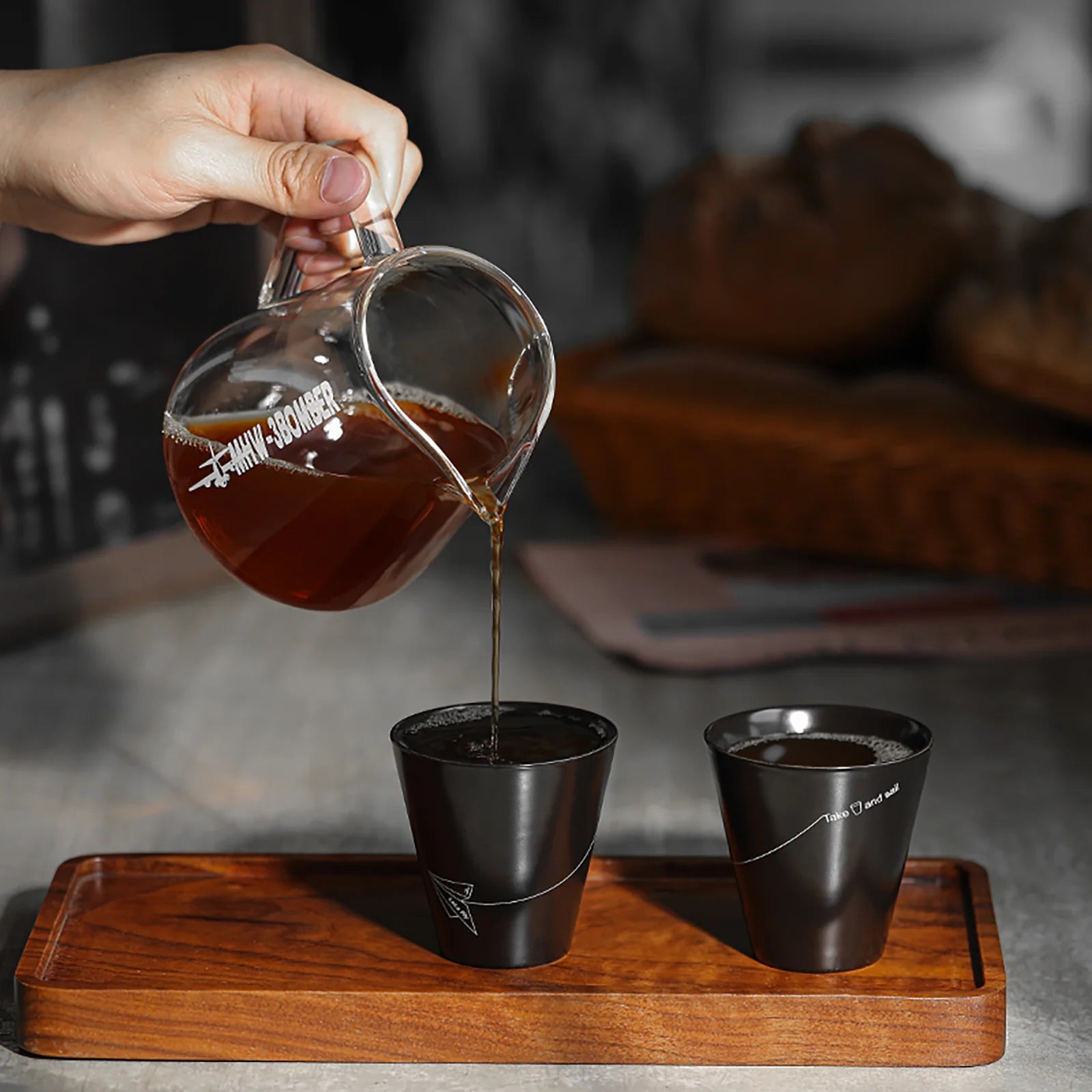Coffee is poured from the MHW-3BOMBER Elf Coffee Server into one of two black cups with fine white lines on a wooden tray. The drip-free design prevents mess as the scene unfolds on a gray table, highlighting the minimalist style of both cups.