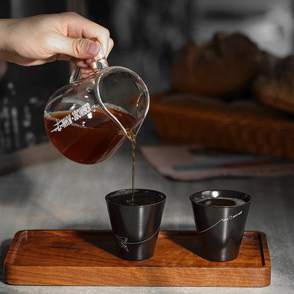 Coffee is poured from the MHW-3BOMBER Elf Coffee Server into one of two black cups with fine white lines on a wooden tray. The drip-free design prevents mess as the scene unfolds on a gray table, highlighting the minimalist style of both cups.