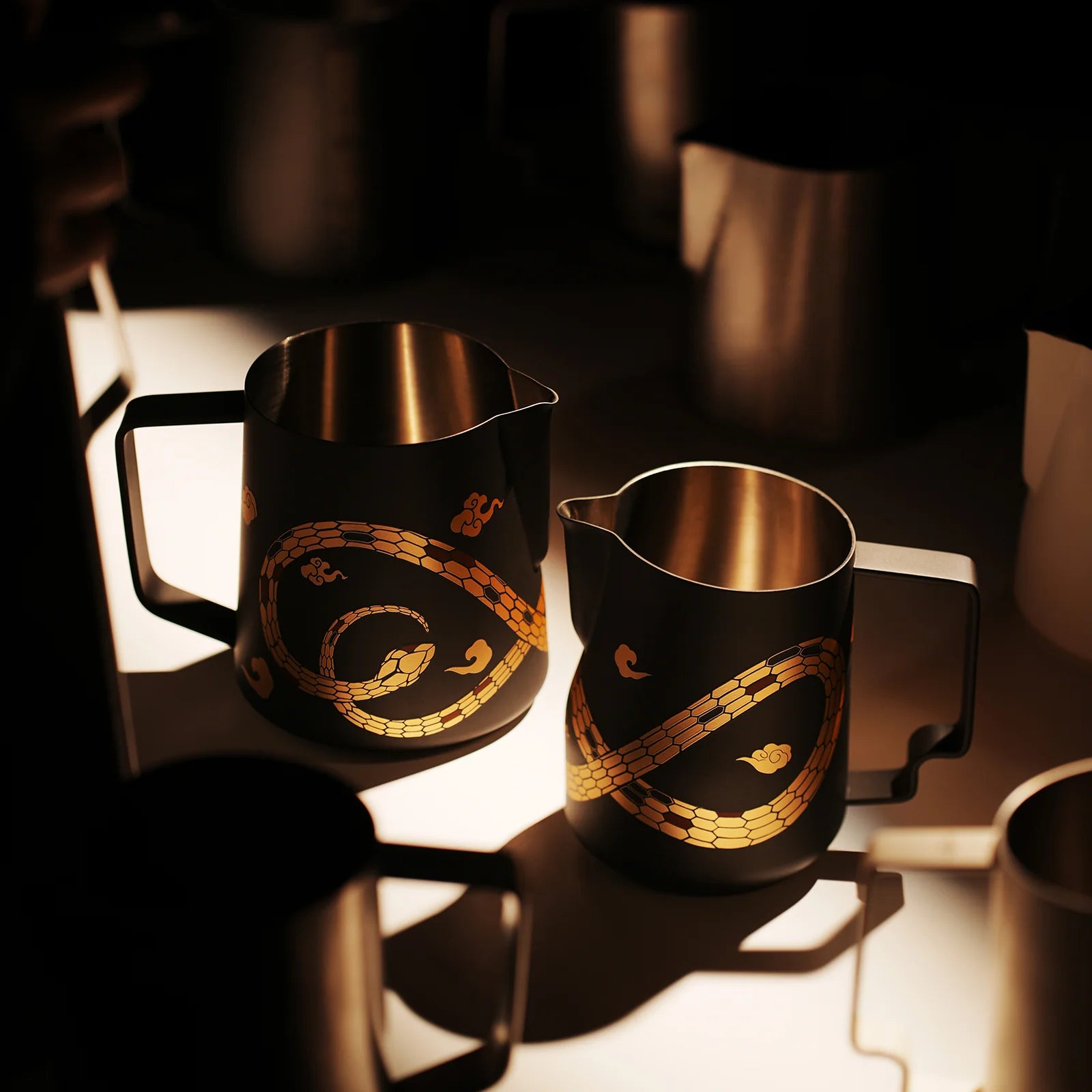 Two black metal MHW-3BOMBER Turbo Milk Pitchers, from the Year of the Snake Limited Edition and crafted from austenitic 304 stainless steel, feature intricate gold snake designs. Soft lighting highlights their glossy finish while other pitchers are partially visible in the dim background.