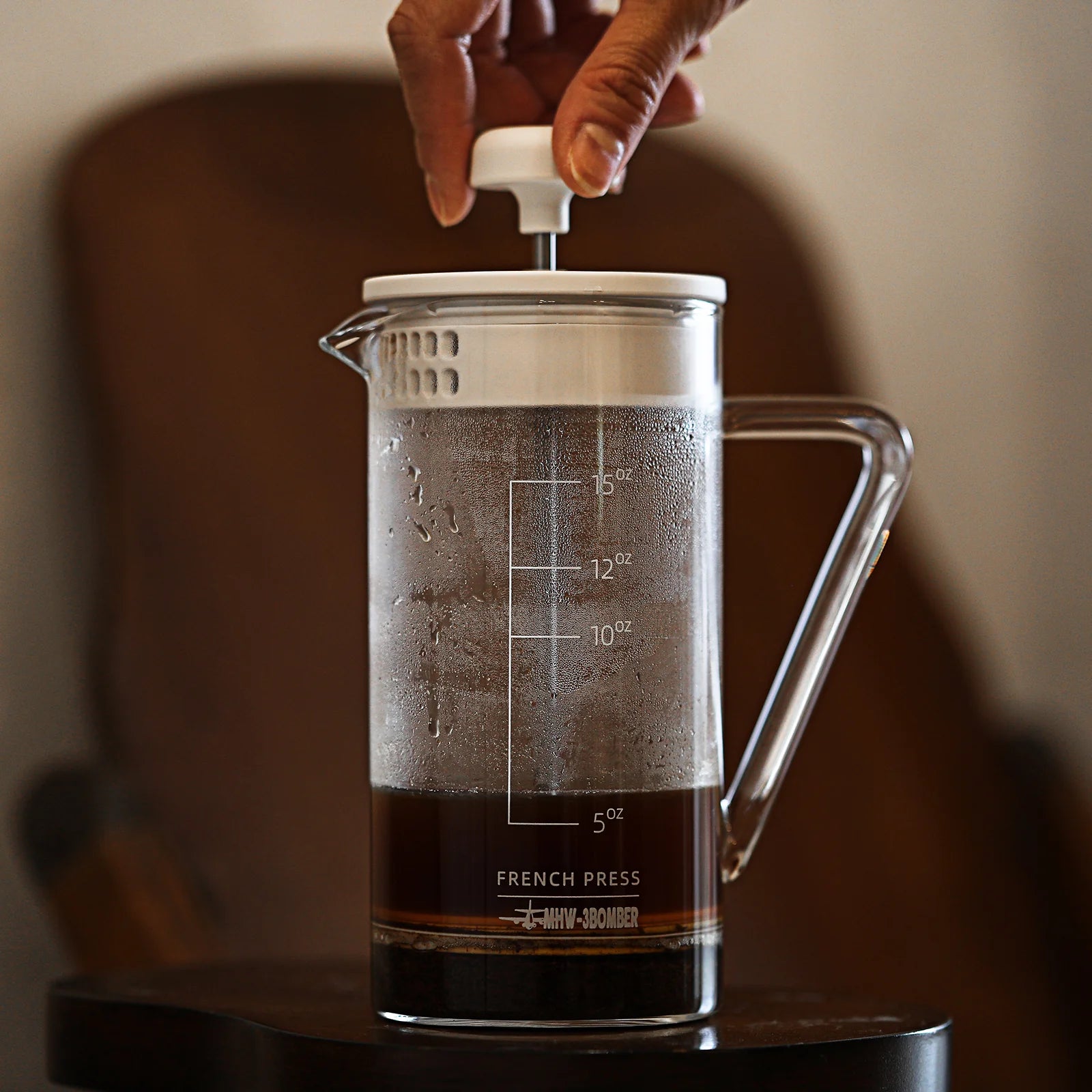 A hand presses the plunger of the MHW-3BOMBER Gimme French Press, highlighting its classic design. The heat-resistant glass shows ounce markers, set against a blurred background with a brown chair.