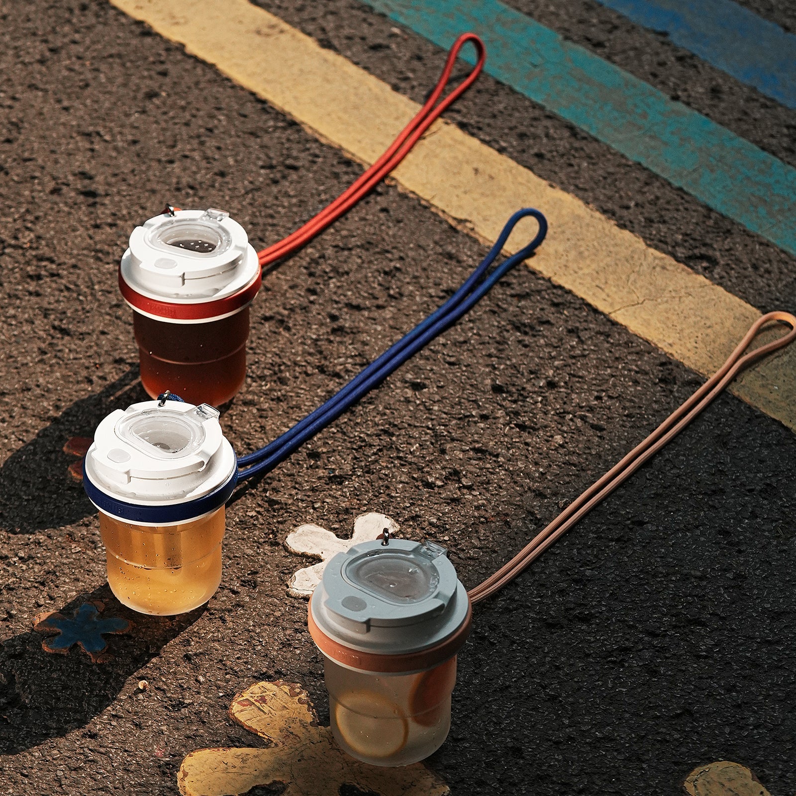 MHW-3BOMBER Cold Brew Cups in red, blue, and tan with straps on pavement. Portable, leak-proof, transparent design.