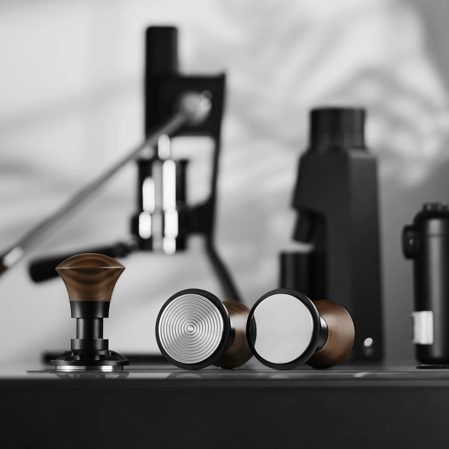 A close-up of the MHW-3BOMBER Armor Series Coffee Tamper 2.0, featuring wooden handles, metallic bases, and a triple spring design for consistent pressure. Espresso machine parts in black are blurred in the background, adding depth and contrast to these innovative coffee tools.