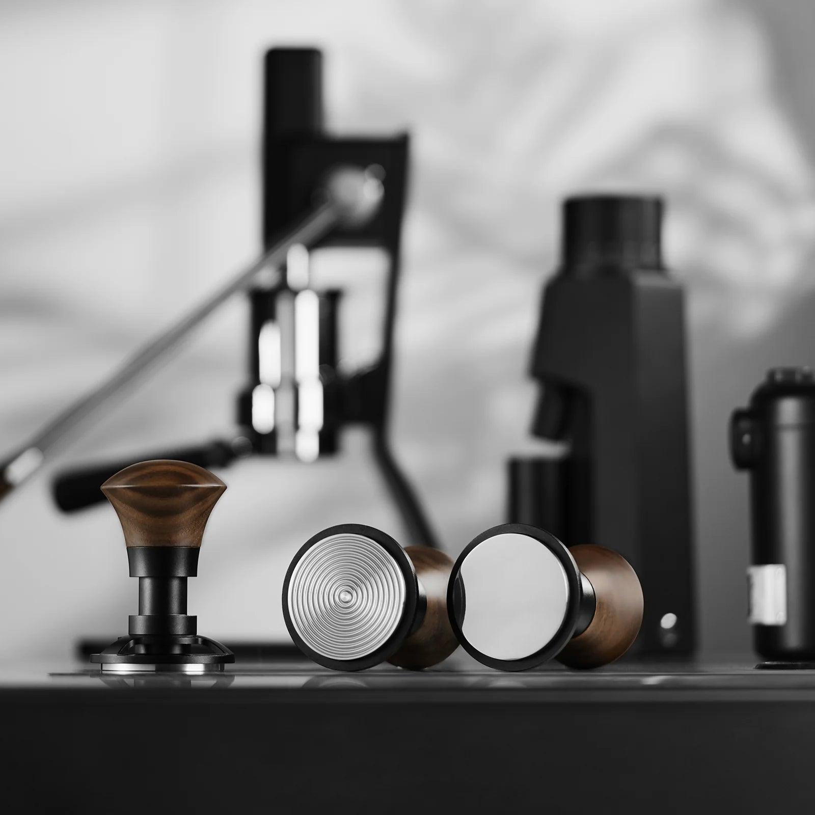 A close-up of the MHW-3BOMBER Armor Series Coffee Tamper 2.0, featuring wooden handles, metallic bases, and a triple spring design for consistent pressure. Espresso machine parts in black are blurred in the background, adding depth and contrast to these innovative coffee tools.