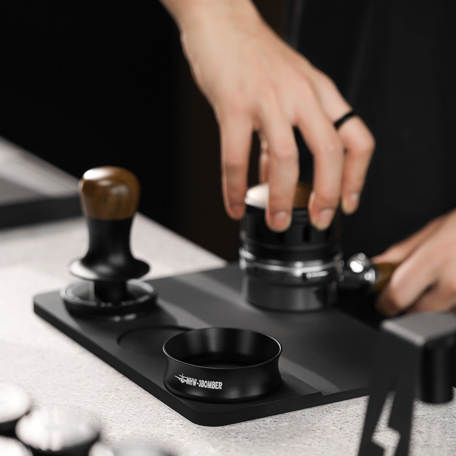 A person expertly uses a coffee tamper on a black mat, surrounded by tools like another tamper and an espresso dosing funnel. The MHW-3BOMBER Espresso Magnetic Coffee Dosing Funnel adds flair to the kitchen or coffee bar setting.