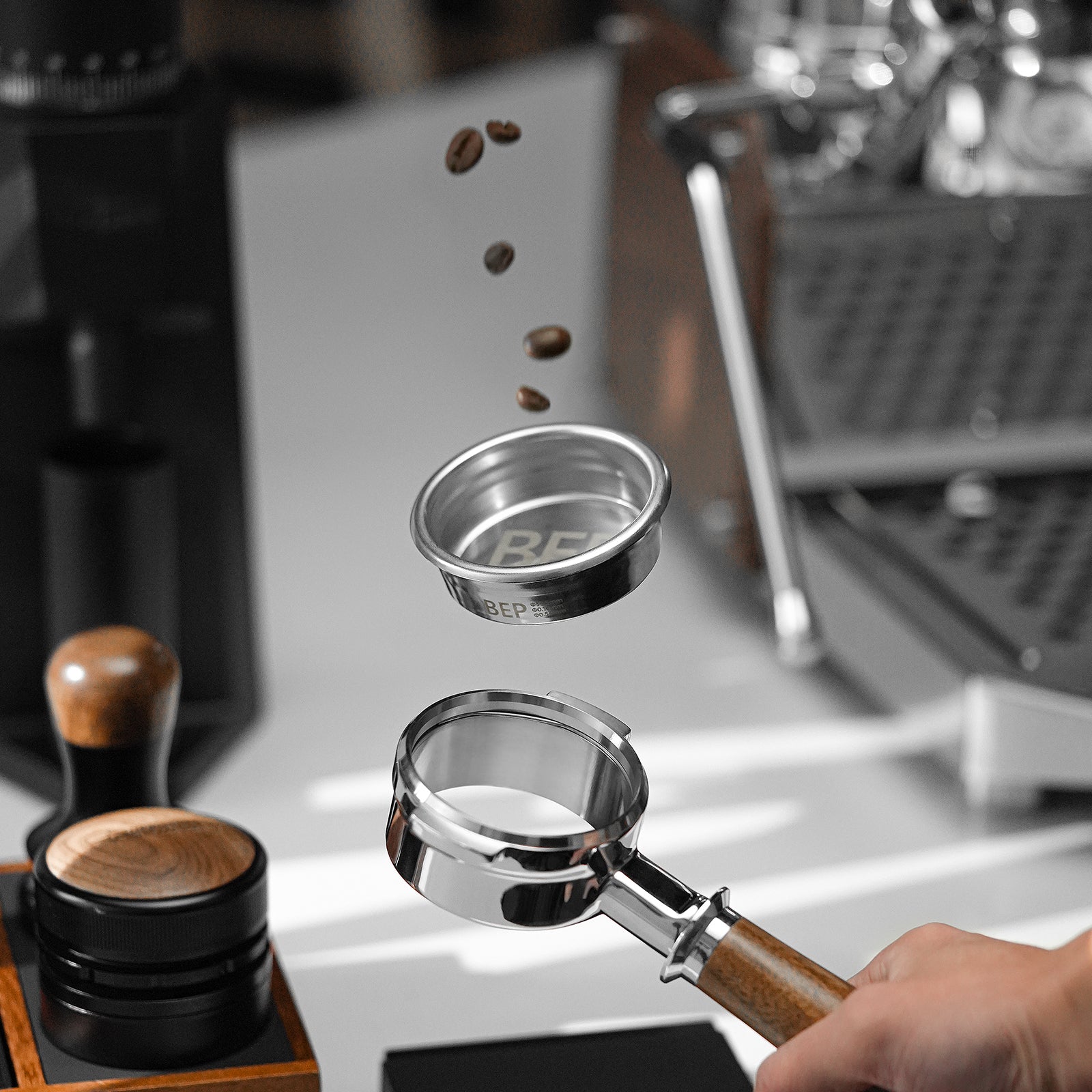 A hand holds the MHW-3BOMBER BEP Butterfly Coffee Filter Basket 2.0 58.5mm under a metal container, with coffee beans suspended mid-air above it. In the background, a wooden tamper and stainless steel espresso machine elegantly blur on a white surface.