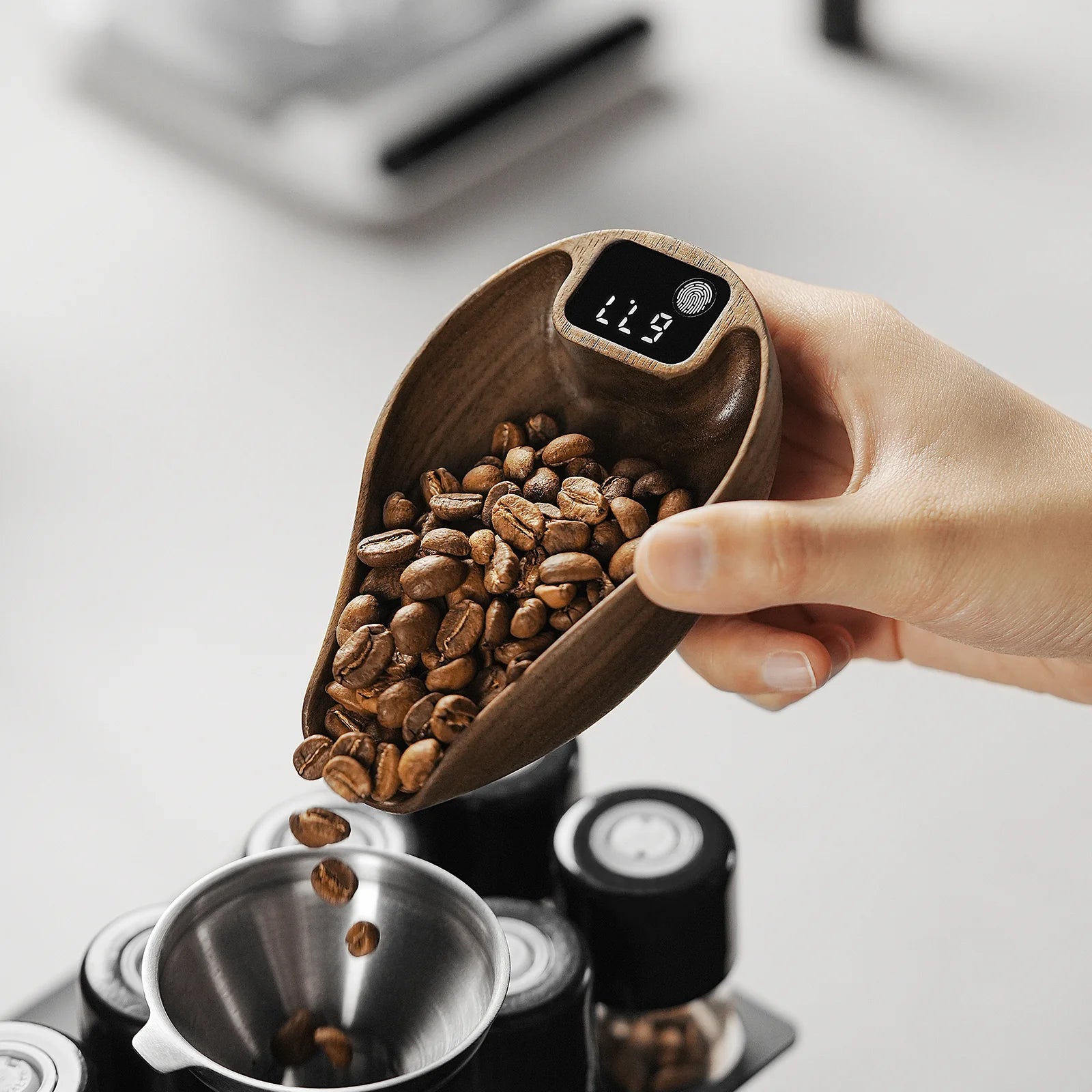 A hand holds a wooden scoop with coffee beans over a metal funnel, displaying the MHW-3BOMBER Ovo Coffee Scale reading 11.7. The blurred background reveals parts of a kitchen setup, illustrating an essential brewing process.