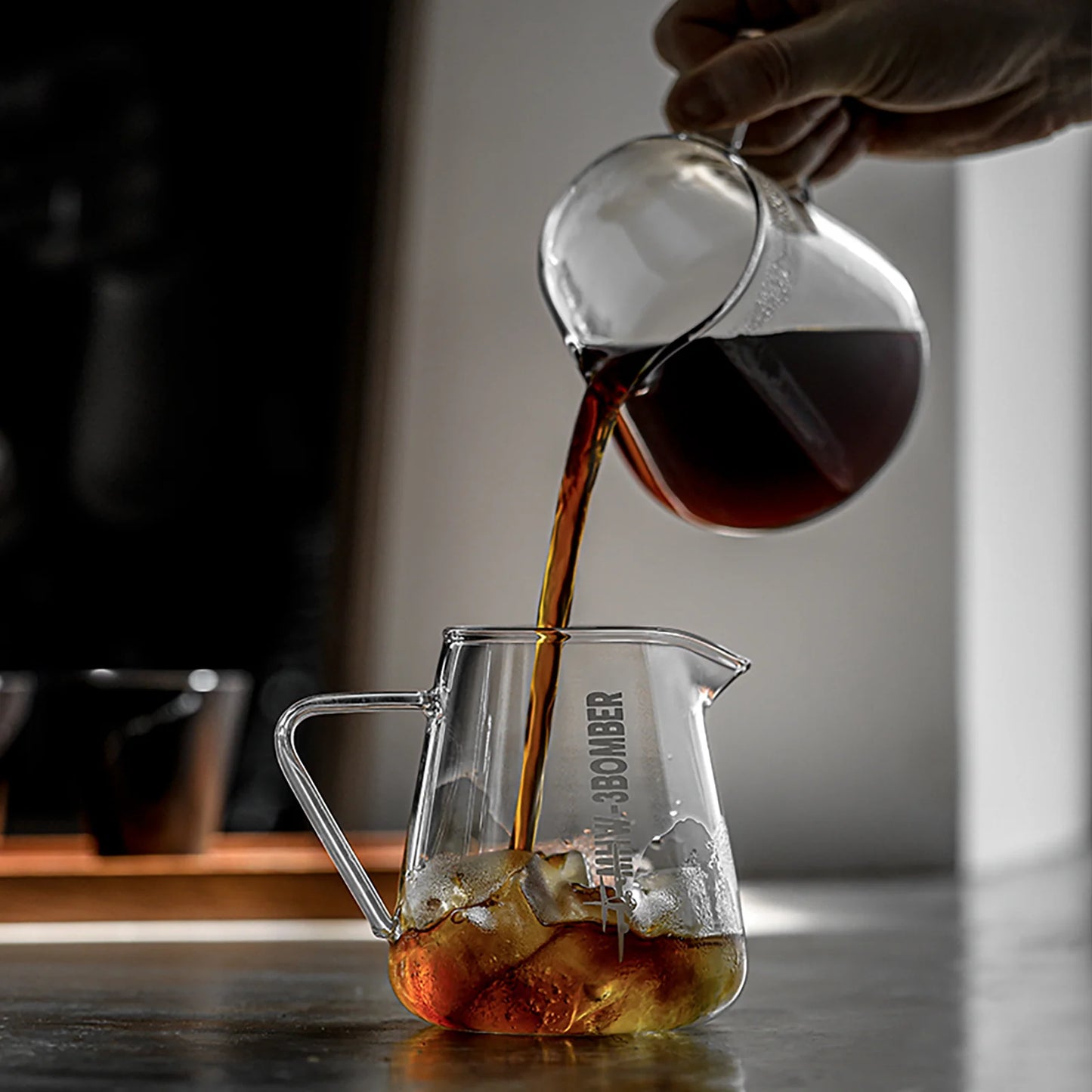 A hand pours coffee from a MHW-3BOMBER Elf Coffee Server, ensuring a drip-free pour into an ice-filled jug on a dark countertop. In the background, light reflects off glass surfaces with a window and two cups blurred.