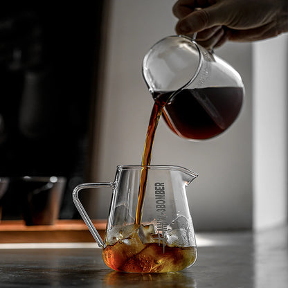 A hand pours coffee from a MHW-3BOMBER Elf Coffee Server, ensuring a drip-free pour into an ice-filled jug on a dark countertop. In the background, light reflects off glass surfaces with a window and two cups blurred.