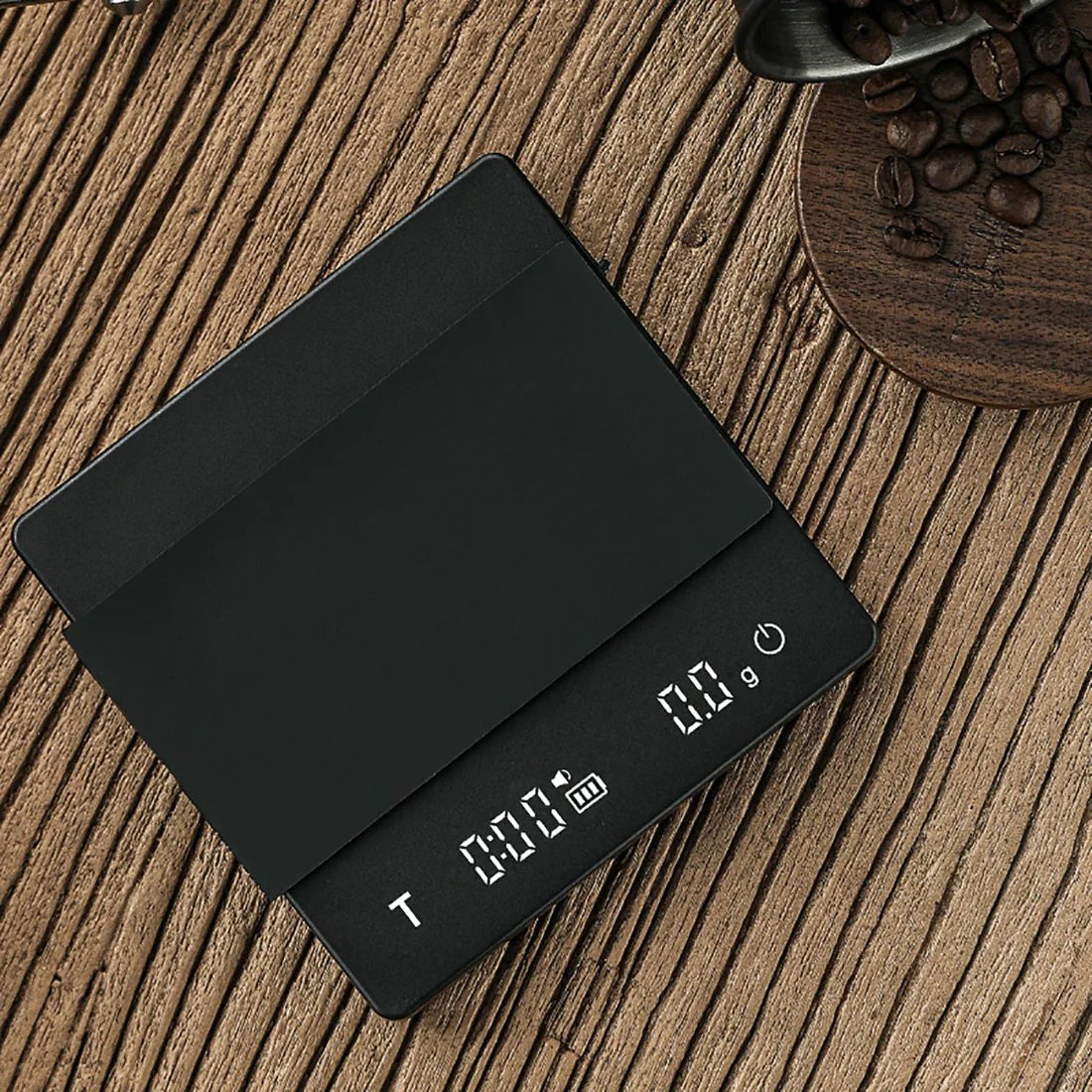 The sleek MHW-3BOMBER Espresso Scale Cube Coffee Scale 2.0, with its high precision and black surface, shows zero grams on a wooden table. Nearby, a wooden coaster holds rich coffee beans with a metal scoop ready to enhance your brewing experience.