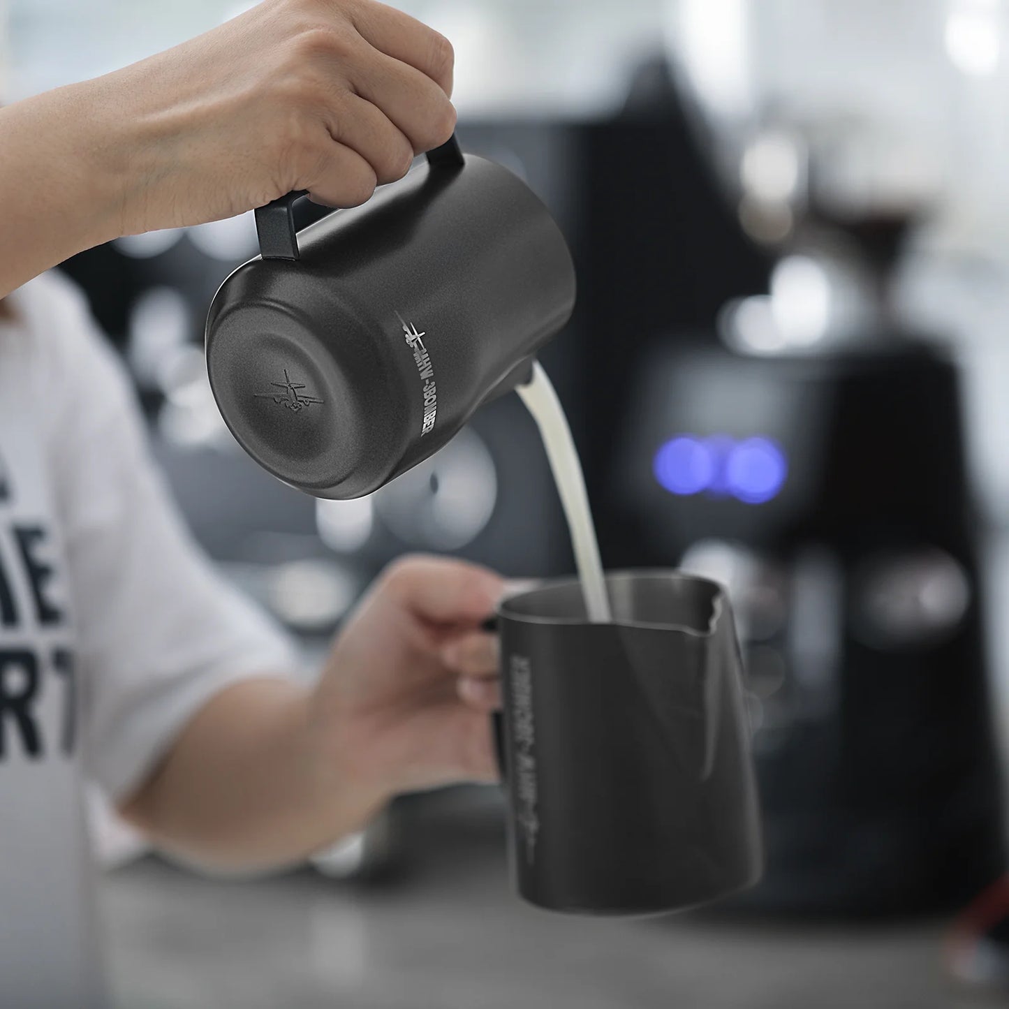 A person expertly pours milk from an MHW-3BOMBER Turbo Milk Pitcher Matte Black into a black cup, with a coffee machine glowing in the blurred background, perfect for latte art. Their shirt partially displays THE ARTS, adding an artistic touch to the scene.