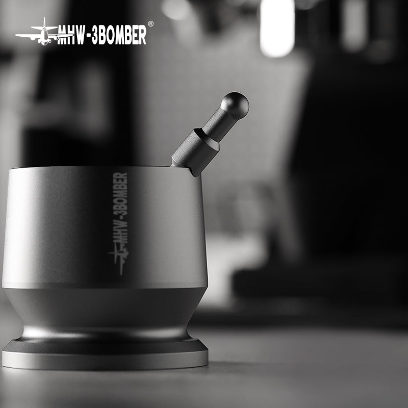 A sleek, modern silver MHW-3BOMBER Espresso Coffee Powder Dosing Funnel with a cylindrical base and lever is shown in close-up. The text MHW-3BOMBER appears on the side, highlighted by a blurred, monochromatic background that emphasizes its coffee brewing potential.