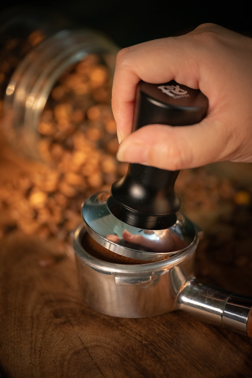 FIX COFFEE tamper with black handle & steel base pressing grounds into portafilter. Constant force for level tamping.