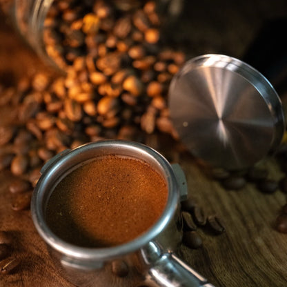 FIX COFFEE tamper with grounds, stainless steel, spring mechanism visible, loose coffee beans on wood. 