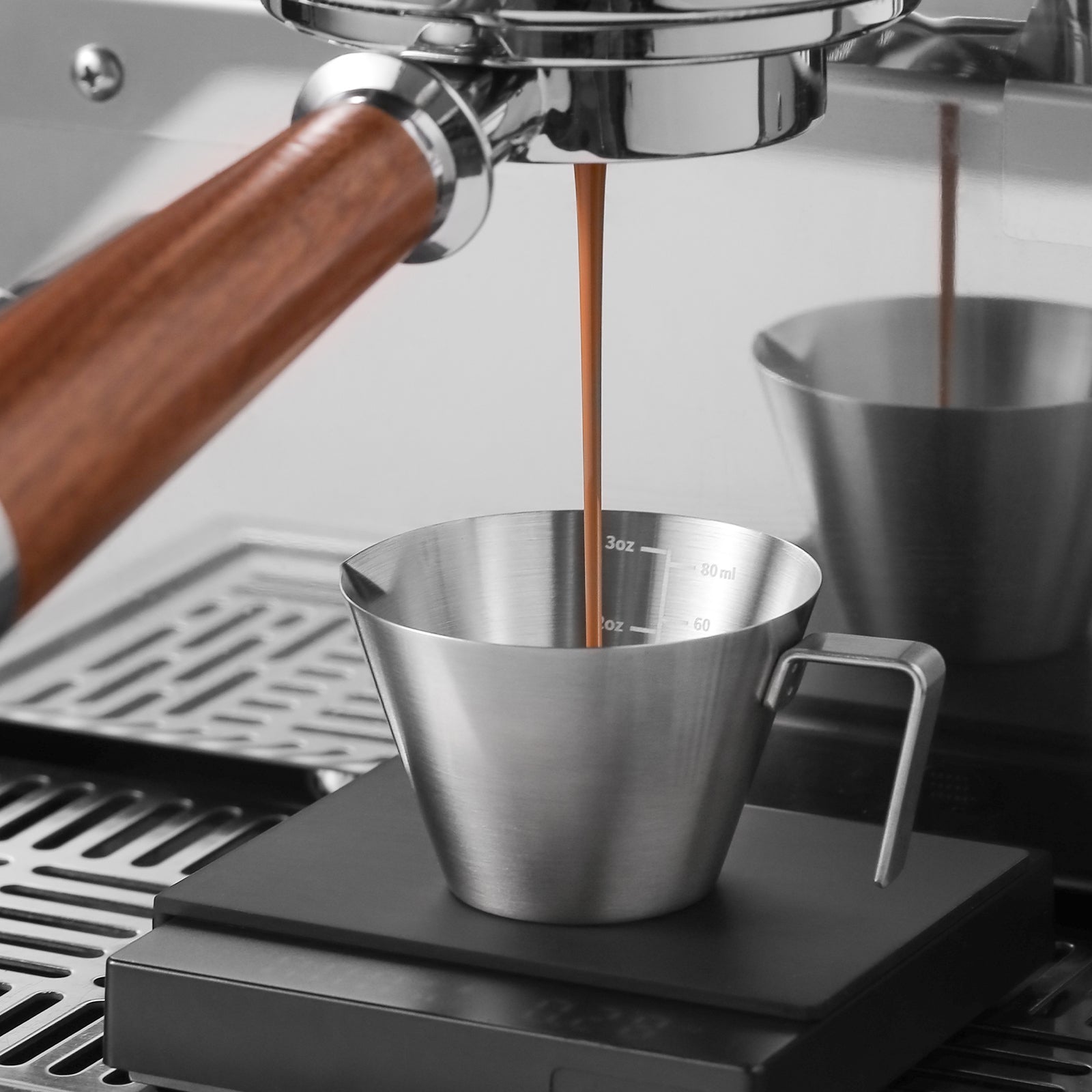 Espresso flows from a coffee machine with a wooden handle into an MHW-3BOMBER Stainless Steel Measuring Cup, featuring volume markings, placed on a digital scale. The setup rests on a textured metallic surface.