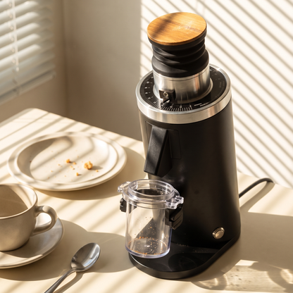 DF54 V4 grinder in matte black with wooden lid on a sunlit table, dosing cup attached, beside a cup of coffee