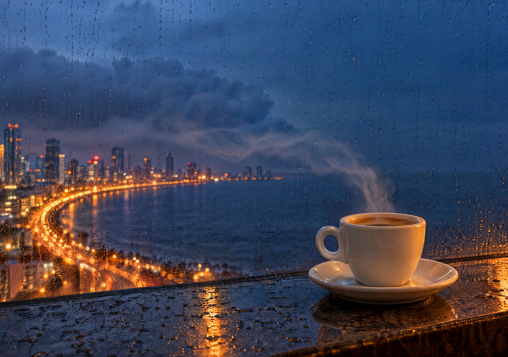 Marine Drive in Mumbai at rainy dusk through rain streaked glass: low monsoon clouds, the curve of golden street lights along the sea, and a steaming espresso cup on the wet ledge on the right. Made with AI.