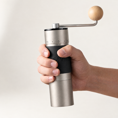 A hand holding the silver KINGrinder K6 by its black grip, showing the complete grinder, steel crank and wooden knob.
