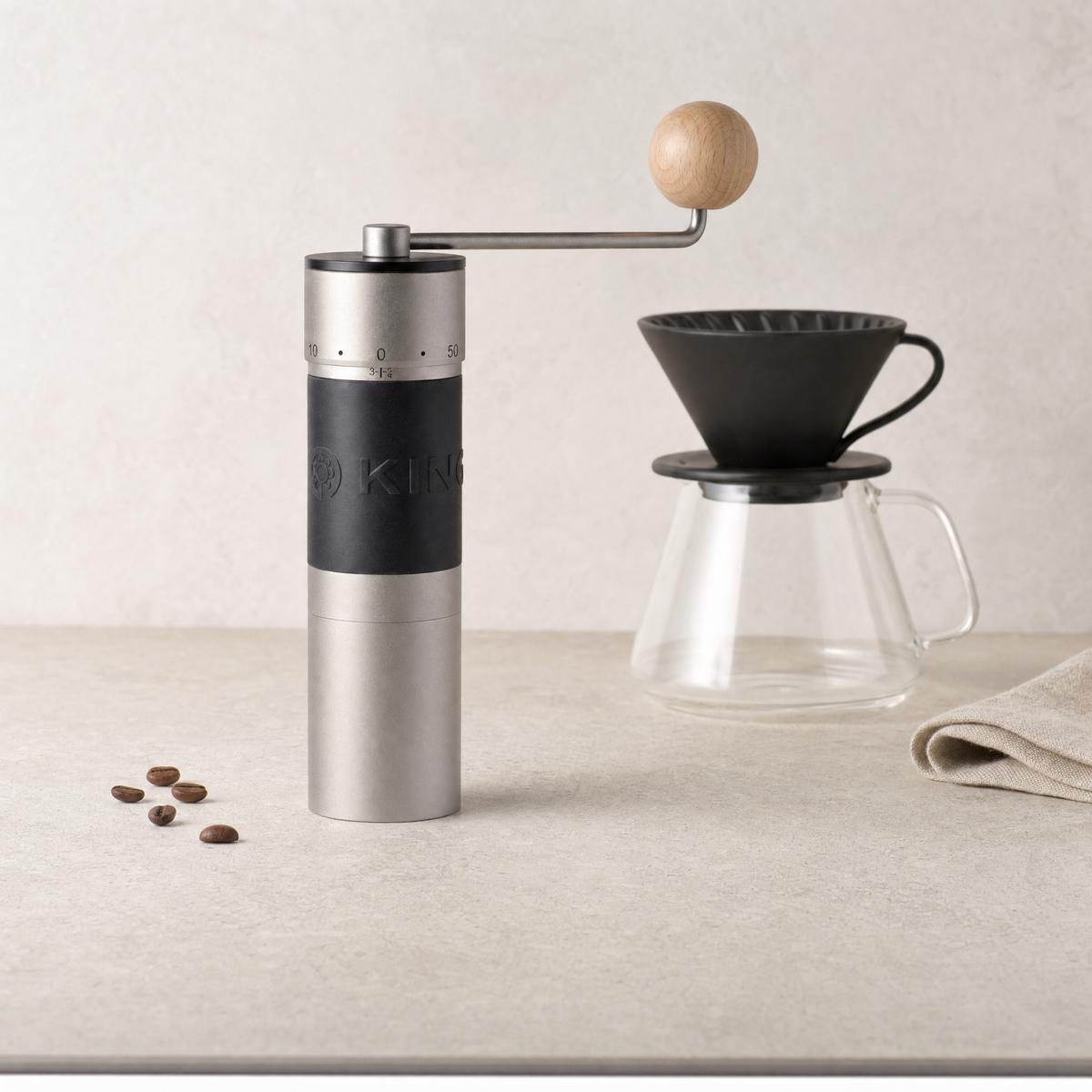 The silver KINGrinder K6 standing upright on a stone counter beside a pour-over brewer and glass server.