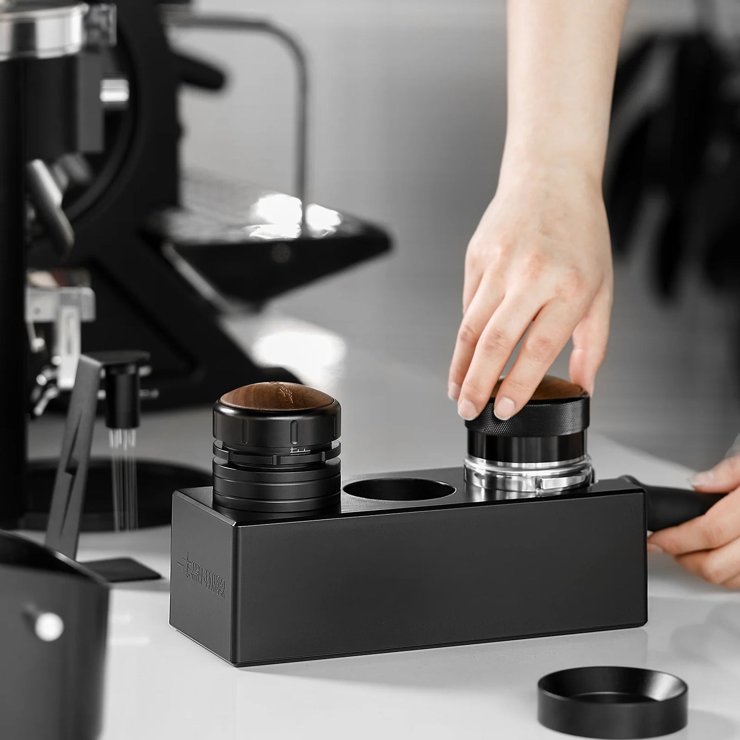 MHW-3BOMBER tamper station. Black ABS portafilter holder. Espresso tools. On counter. Hands visible.