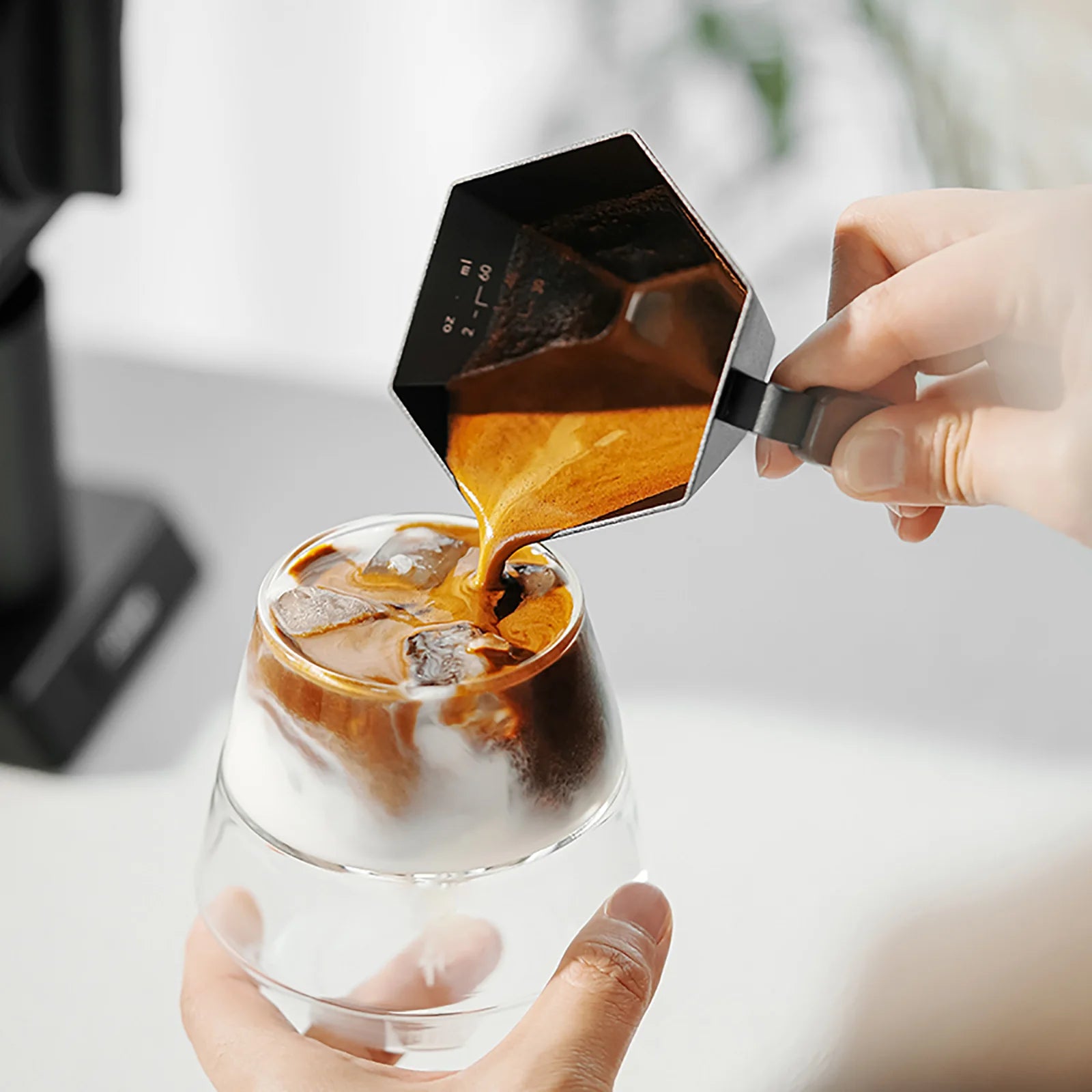 MHW-3BOMBER cup pouring espresso into ice-filled glass. Black, hexagonal design, open wood handle. Precise coffee brewing.